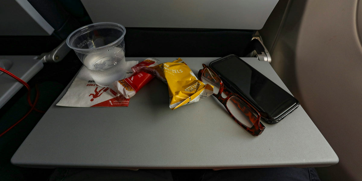 Airplane tray table with a plastic cup of water, snack packets, reading glasses and a phone