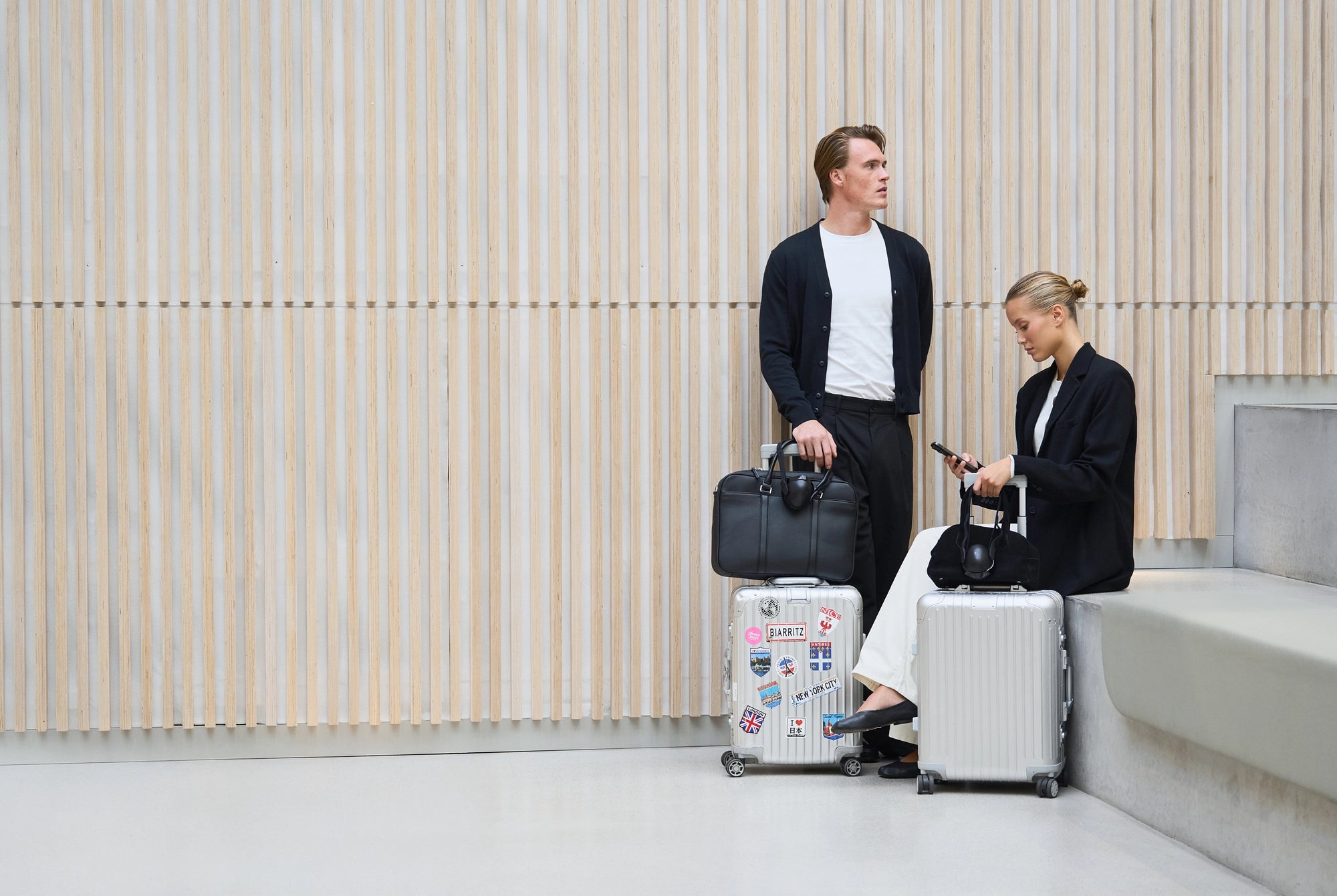 Two people with luggage standing against a light-colored wall.