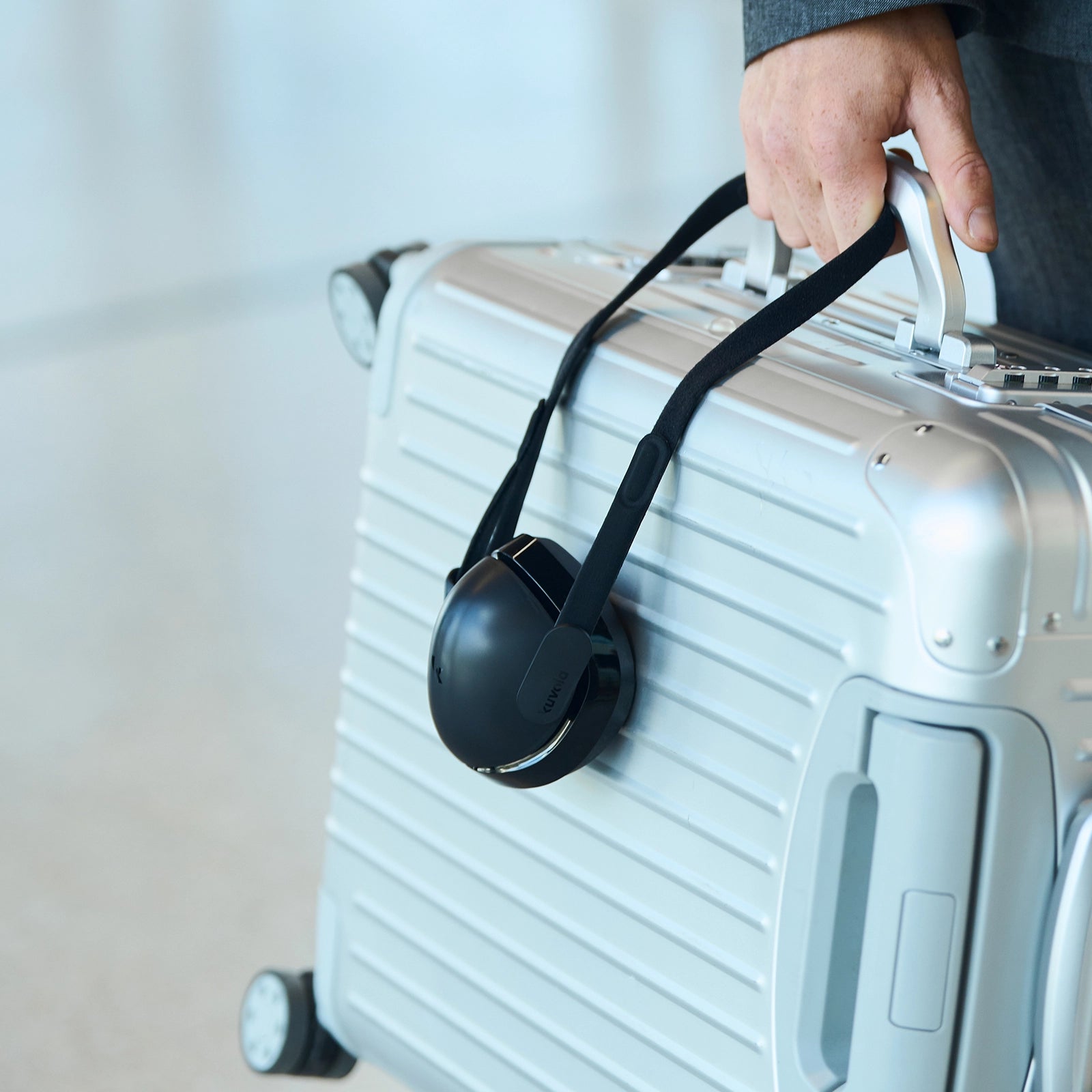 Person holding a silver suitcase with a black luggage strap against a light background